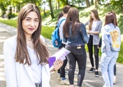 Studentka trzymająca w ręku książkę uśmiecha się do zdjęcia. W tle stoi grupa studentów. Znajdują się w parku.