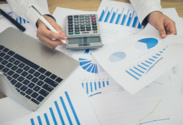 Dwie dłonie opierają się na stole. W jednej ktoś trzyma długopis, w drugiej kartkę z wykresami. Obok leży więcej kartek z wykresami i laptop.