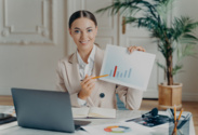 Kobieta trzymająca długopis i kartkę z wykresem słupkowym. Wskazuje na słupek. Siedzi przy biurku, na którym leży kartka i laptop.