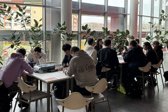 Grupa kilkunastu osób. Siedzą przy dwóch stołach. Grają w gry planszowe.